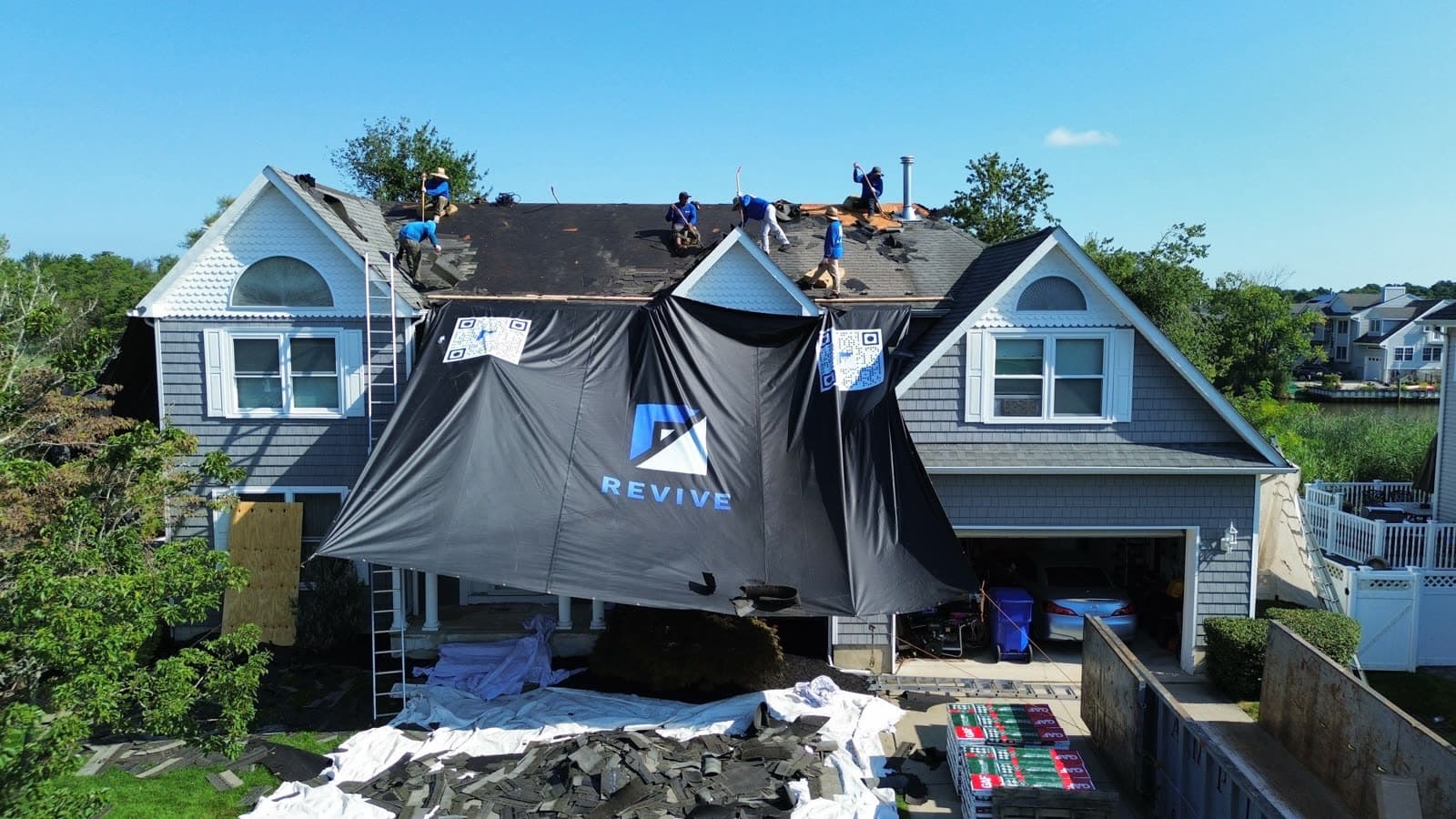 Revive crew tearing off and replacing a roof on a New Jersey coastal home, with the Revive-branded debris tarp protecting the siding below